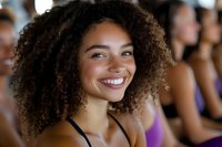 Happy young mixed-race woman with curly hair smiling during fitness class or group workout session in gym with blurred people in background.