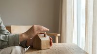 Woman hand placing coin into wooden house-shaped piggy bank on table near window. Saving money for home purchase, mortgage payment, real estate investment concept.