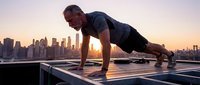 Athletic man performing plank exercise on rooftop at sunrise with city skyline backdrop. Fitness training and strength workout concept.