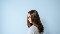 Young woman with long brown hair and fair complexion, wearing casual gray clothing, positioned in profile against a light blue background for portrait and lifestyle use.