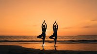 Two women in silhouette practice yoga on a beach at sunset, standing in tree pose with arms raised overhead. Golden hour light creates a serene wellness and fitness scene.