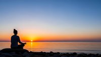 Person meditating in lotus position on rocky shore during golden sunrise over calm ocean waters creating peaceful zen atmosphere for wellness content.