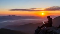 Young man with laptop working remotely on mountain peak during golden sunrise with layered hills and valleys below for digital nomad lifestyle content.