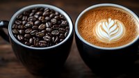 Fresh roasted coffee beans in black ceramic cup next to creamy latte with artistic leaf pattern foam art on rustic wooden table background, moody lighting.