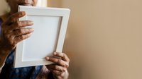 Mature adult with dark skin tone holding empty white wooden frame against neutral beige wall, suitable for framing artwork, photos, or displaying custom designs and home decor projects.