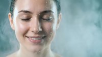 Young woman with water droplets on face enjoying refreshing shower or spa treatment with closed eyes and gentle smile against soft background.