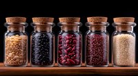 Various dried legumes and grains in glass jars with cork stoppers arranged in row on wooden surface against dark background, showing natural food ingredients.