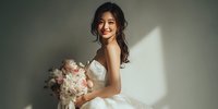 Young Asian bride in elegant white wedding dress holding pink and white flower bouquet, smiling joyfully in soft natural light for bridal photography.