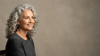 Mature woman with long gray curly hair wearing dark sweater, smiling at camera against neutral beige background. Portrait for advertising, healthcare, lifestyle.
