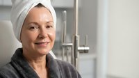 Mature woman with towel on head smiling in modern bathroom after shower wearing gray bathrobe enjoying morning skincare routine and wellness lifestyle.