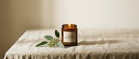 Amber glass candle with blank label on linen fabric with fresh green foliage and white flowers, ideal for home decor, wellness, and product branding designs.