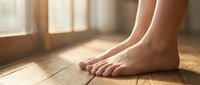 Bare feet of a young woman standing on warm wooden floor in soft morning sunlight, showcasing healthy skin and natural beauty.