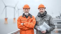 Senior male engineers in orange hard hats at wind farm, one wearing orange safety jacket with arms crossed, other in gray coat holding documents, wind turbines in background.