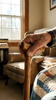 Senior man's weathered hand gripping wooden cane in cozy bedroom with armchair, books, and natural window light. Depicts aging, mobility assistance, retirement living, healthcare support.