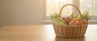 Wicker basket filled with fresh vegetables including carrots, cucumbers and herbs on wooden table by window with natural sunlight for farmers market or grocery shopping concepts.