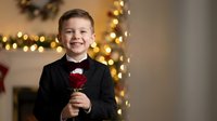 Young Caucasian boy in formal black suit holding red rose with warm Christmas lights bokeh background creating festive holiday atmosphere for celebrations.