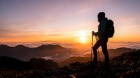 Silhouetted hiker with backpack and trekking poles stands on mountain summit at golden sunrise, gazing at misty valleys and distant peaks below, capturing adventure and exploration for travel and outdoor lifestyle content.