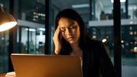 Stressed Asian businesswoman working late at night in modern office, experiencing headache and fatigue while using laptop computer for overtime work.