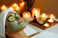 Woman receiving facial spa treatment with cucumber slices and green clay mask, relaxing in candlelit wellness setting for skincare, beauty and self-care concepts.