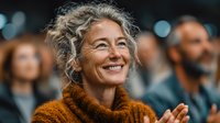 Mature woman with gray curly hair smiling warmly at conference or seminar, wearing orange sweater, audience blurred in background for business content.