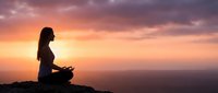 Woman silhouette meditating in lotus pose on mountain peak during golden sunset with dramatic colorful sky. Peaceful mindfulness and spiritual wellness concept.