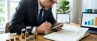 Senior businessman analyzing financial data on smartphone while reviewing investment charts and coin stacks in modern office environment.