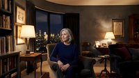 Professional middle-aged Caucasian businesswoman in navy sweater sitting confidently in upscale office with city skyline view at night.