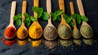 Colorful spices and herbs in wooden spoons on dark slate background. Fresh mint leaves accent aromatic seasonings for cooking and culinary preparation.