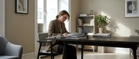 Young adult male with light skin tone, wearing casual brown shirt, focused on work at black desk in modern home office with natural lighting and minimalist decor.
