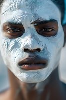 Close-up portrait of African American woman with white clay facial mask, showing intense gaze and natural lips against blue background. Wellness and skincare concept.