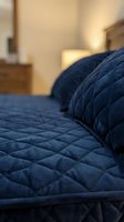 Close-up of navy quilted bedding with pillows on bed in warm-lit bedroom. Soft focus background shows nightstand and furniture. Use for home decor, bedroom design, hospitality, and interior styling projects.