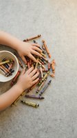 Child hands reaching for colorful crayons scattered on concrete floor from white bowl, creative art supplies for drawing and coloring activities.