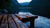 Wooden pier planks at mountain lake during sunset, dramatic moody atmosphere with calm water surface reflecting evening light and distant peaks.