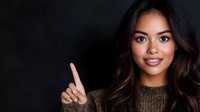 Young Asian woman with long dark wavy hair and bright smile pointing finger up against black background, wearing brown knit sweater. Professional portrait with copy space.