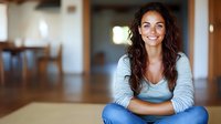 Young Hispanic woman with long dark wavy hair wearing casual grey top and jeans sits cross-legged on floor, displaying genuine warm smile in modern interior setting.