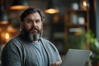Middle-aged Caucasian man with beard working on laptop in modern office space with warm lighting and blurred background for business concepts.