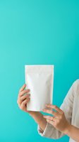 Woman holding blank white stand-up pouch against turquoise background for product packaging mockup, branding, and retail display purposes.