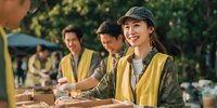 Young Asian woman volunteer in yellow vest and cap smiling while distributing food at outdoor community service event with diverse team members.