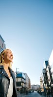 Young woman with blonde hair, Caucasian ethnicity, wearing black leather jacket and white top, smiling while looking upward in urban street setting with clear blue sky and modern buildings.