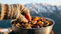 Hiker in knit sweater holding bowl of mixed nuts and dried fruits with snowy mountain landscape in background, outdoor snacking and trail food concept.