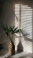 Sunlight through window blinds casting dramatic shadows on white wall with palm plant in striped ceramic vase, creating minimalist morning atmosphere.