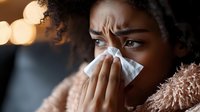 Young African American woman blowing nose into tissue, showing signs of illness while wearing cozy pink sweater against blurred bokeh background.