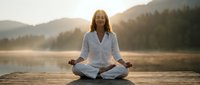 Mature woman meditating in lotus pose on wooden dock at sunrise with mountains and lake in background, peaceful morning wellness retreat.