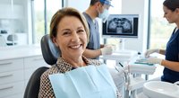 Middle-aged Caucasian woman smiling in dental chair during checkup at modern clinic with dentist and assistant working in background.