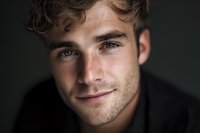 Young handsome Caucasian man with curly brown hair and freckles smiling warmly at camera in close-up portrait against dark background for lifestyle content.
