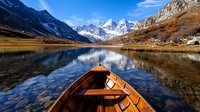 Wooden canoe on pristine alpine lake with snow-capped mountain peaks reflecting in calm water during autumn season in wilderness landscape.