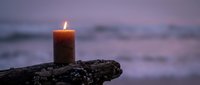 Burning candle on driftwood at twilight beach with soft purple sky and blurred ocean waves creating peaceful meditation atmosphere for relaxation and mindfulness.