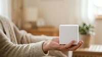Close-up of mature hand holding white cube product mockup in warm home interior with blurred background, suitable for packaging design and product presentation.