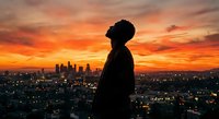 Silhouette of young African American man against dramatic orange sunset sky overlooking illuminated city skyline at dusk for urban lifestyle concepts.
