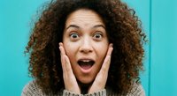Shocked surprised young African American woman with curly hair expressing amazement with hands on face against turquoise background for emotional reactions.